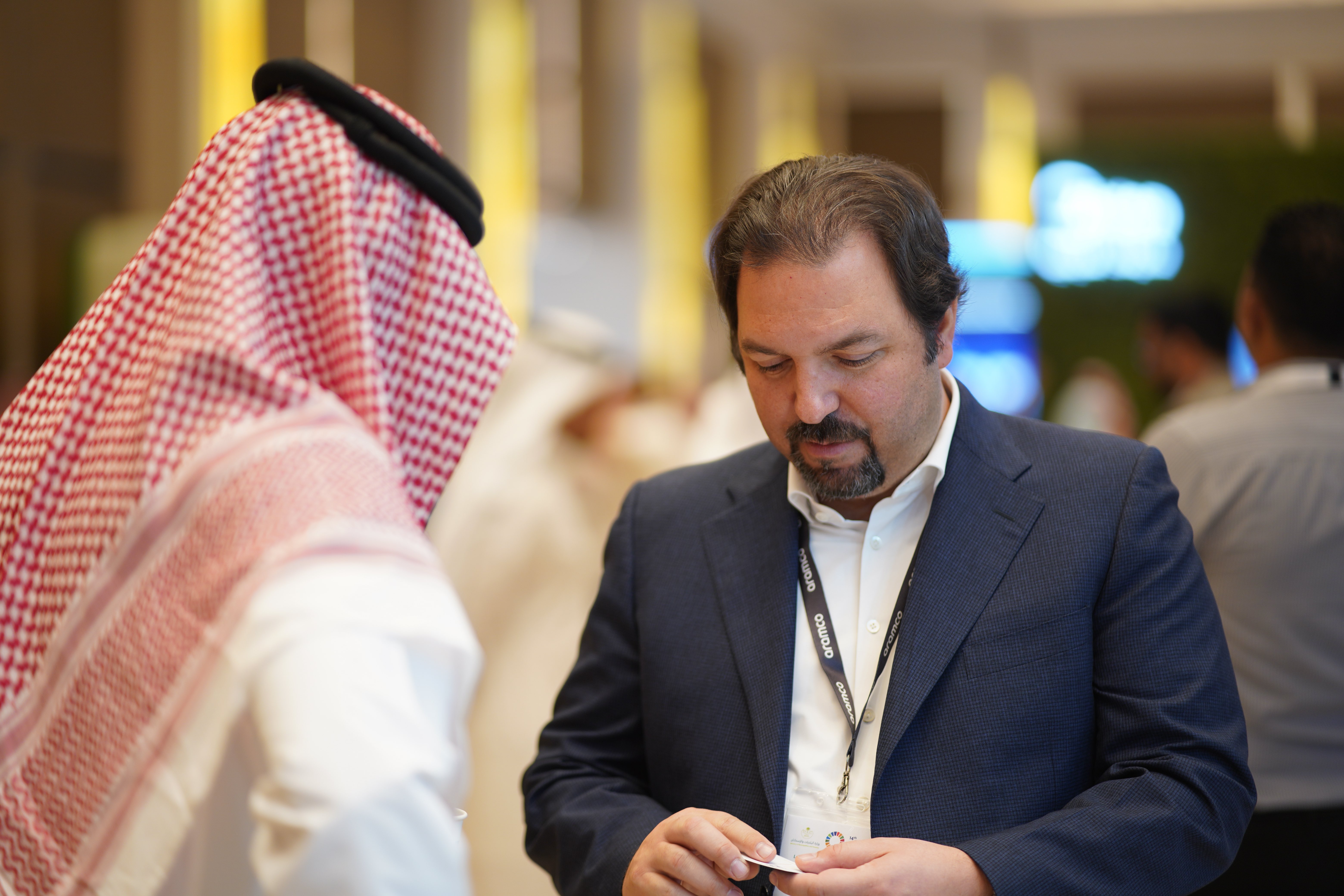 Man in a blue suit looking down at an item in his hands, with another man in traditional Middle Eastern attire standing nearby.